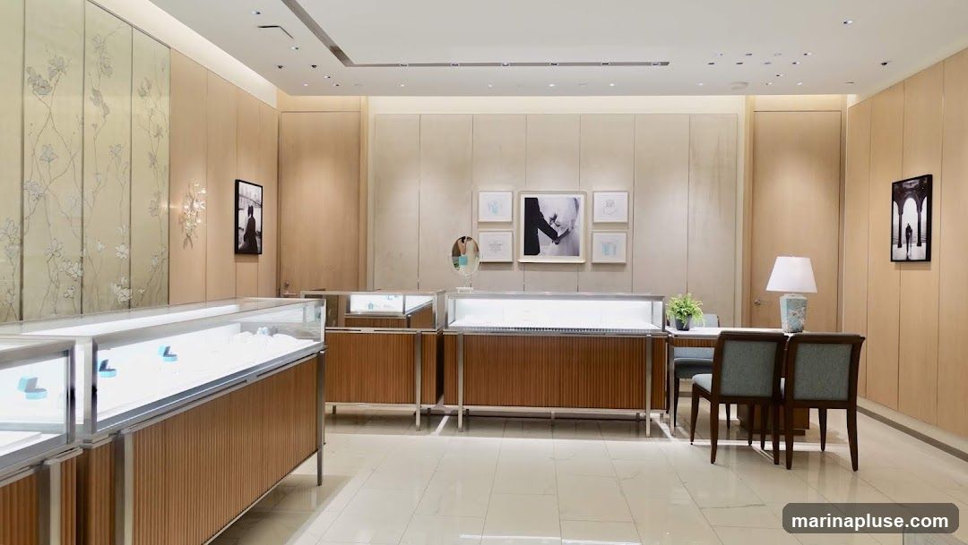 A well-lit jewelry store interior with polished floors and light wood paneling. Display cases with jewelry are visible, along with framed artwork on the walls and a seating area with a lamp.