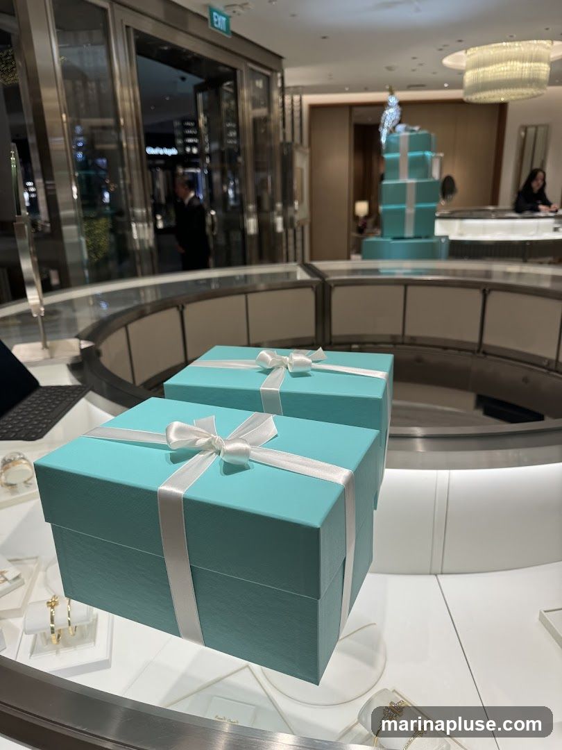 Two iconic Tiffany blue boxes with white ribbon are prominently displayed in the foreground on a glass counter. In the background, the store interior is visible, featuring display cases with jewelry, a tiered display of gift boxes, and a checkout counter where a staff member is assisting a customer. The overall ambiance is luxurious and elegant, typical of a high-end jewelry store.