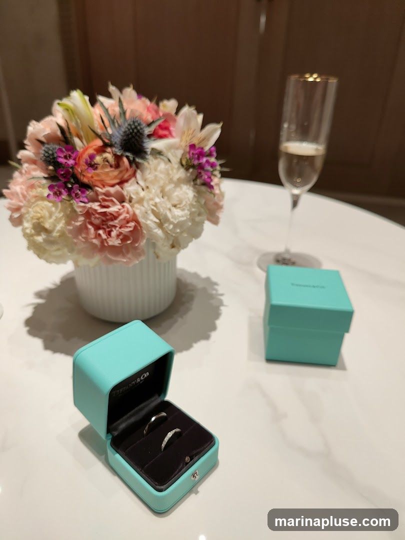 A close-up shot features two engagement rings nestled in an open Tiffany & Co. box. Adjacent to the box is another closed Tiffany & Co. box. A champagne flute filled with bubbly liquid stands to the right, and a bouquet of pink and white flowers in a white vase is on the left. The items are arranged on a white marble surface.