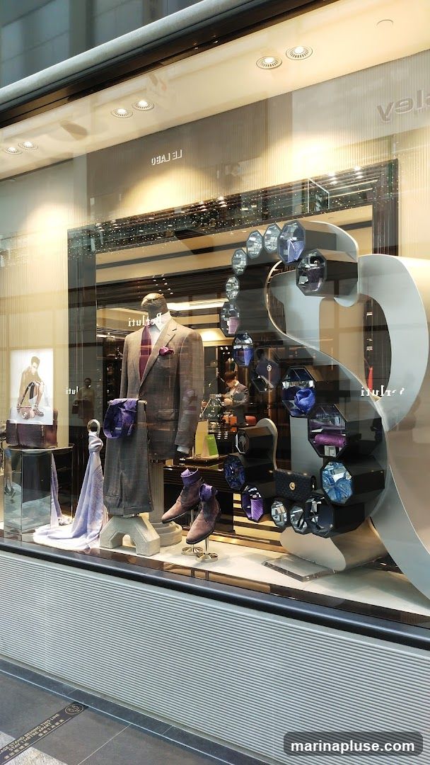 A store window display features a mannequin dressed in a grey suit with a burgundy tie and pocket square. The display also includes a selection of ties, scarves, and shoes. A sign with the text "LE LABO" is visible in the reflection.