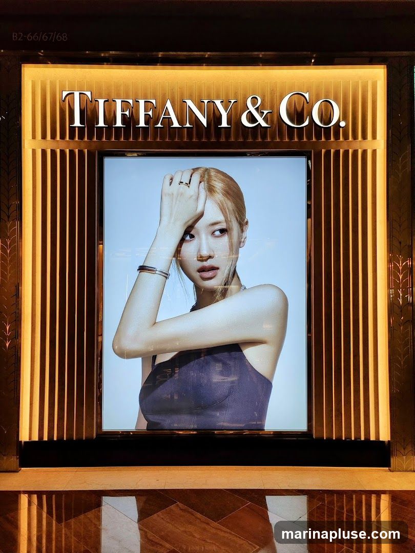 A large illuminated advertisement for Tiffany & Co. features a portrait of a woman with blonde hair. She is wearing a dark top and jewelry, including rings and a bracelet. The Tiffany & Co. logo is prominently displayed above the image. The advertisement is framed by vertical golden slats.