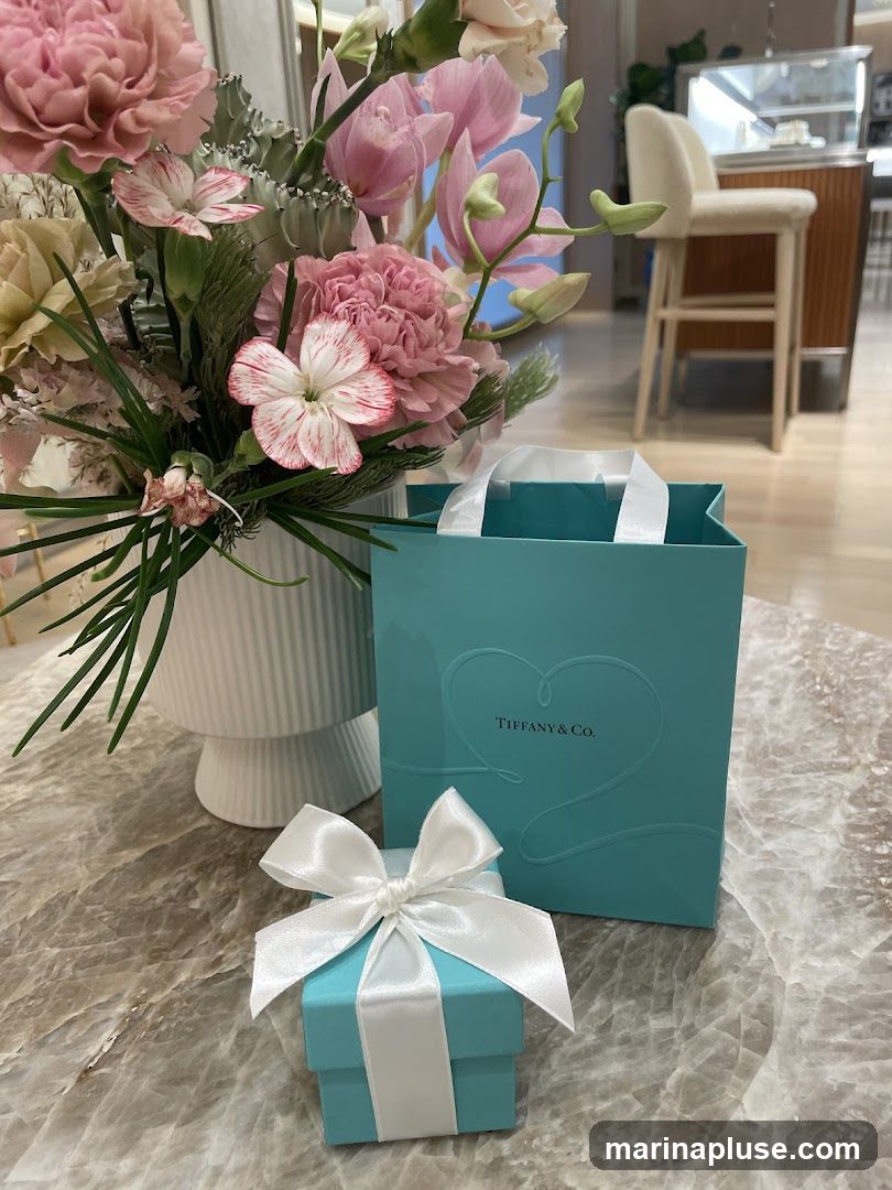 A small blue Tiffany & Co. gift box tied with a white satin ribbon sits on a marble surface next to a larger blue Tiffany & Co. shopping bag. A vase of pink and white flowers is to the left of the box and bag. In the background, a counter with stools suggests a retail environment.
