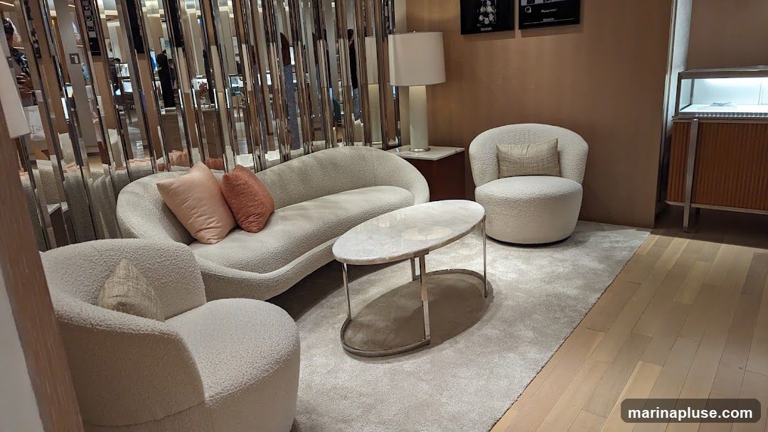 A plush, cream-colored sofa and two armchairs are arranged around a marble-topped coffee table in a well-lit, elegant space. The room features a mirrored wall that reflects the interior, creating a sense of depth and sophistication. A tall lamp stands on a side table, and a display cabinet with illuminated shelves is visible in the background. The flooring transitions from a soft carpet to polished wood.