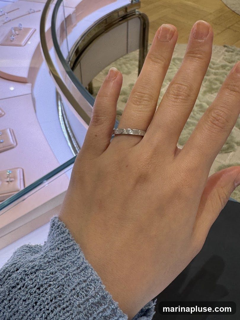 A close-up shot of a woman's left hand, adorned with a silver ring featuring small diamonds and engraved text. The hand is positioned in front of a display case in what appears to be a jewelry store. The display case, with its curved glass and illuminated shelves, showcases various jewelry items. The background is softly blurred, suggesting an indoor retail environment. The woman is wearing a light blue, textured sweater.