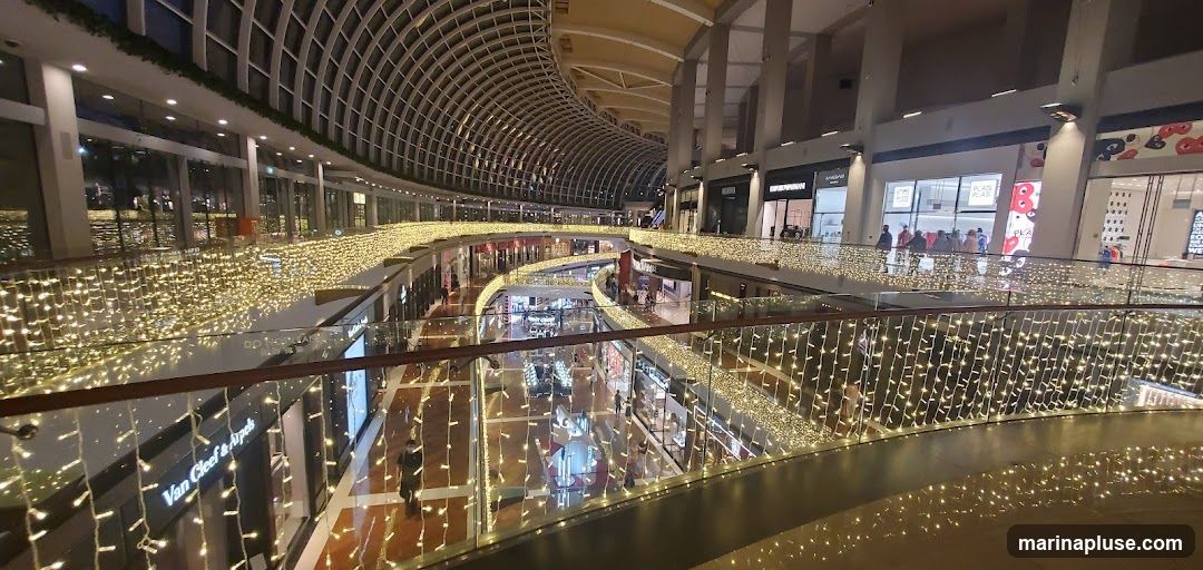 The image shows the interior of a multi-level shopping mall, adorned with numerous strings of warm white fairy lights along the railings and architectural elements. The mall features a distinctive curved ceiling structure. Shops are visible on different levels, with some storefronts identifiable, such as 'Van Cleef & Arpels'. People are seen browsing and walking through the mall, suggesting it is open for business. The overall ambiance is festive and illuminated.