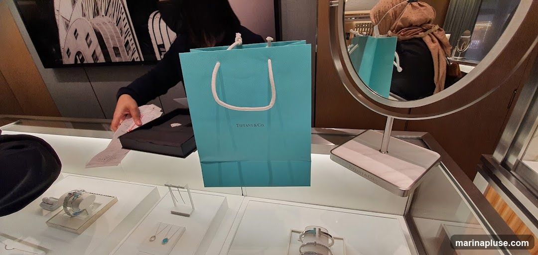 A Tiffany & Co. shopping bag and a black gift box are on display in a jewelry store. A person's hand is seen reaching for receipts. The display cases show various jewelry items, including watches and necklaces. A mirror reflects a person wearing a headscarf.