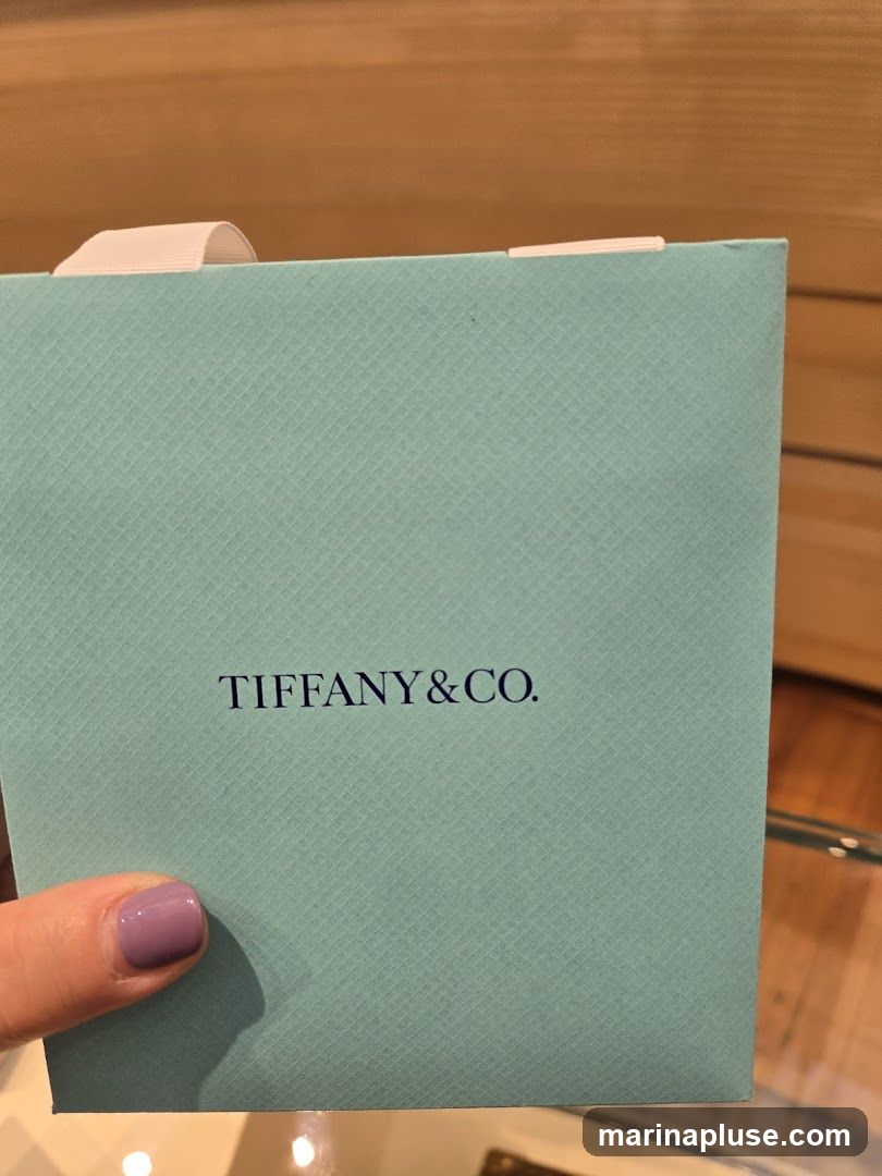 A hand with purple nail polish holds a light blue Tiffany & Co. box. The box has a textured surface and a white ribbon tied around the top. The Tiffany & Co. logo is printed in dark blue on the front of the box.