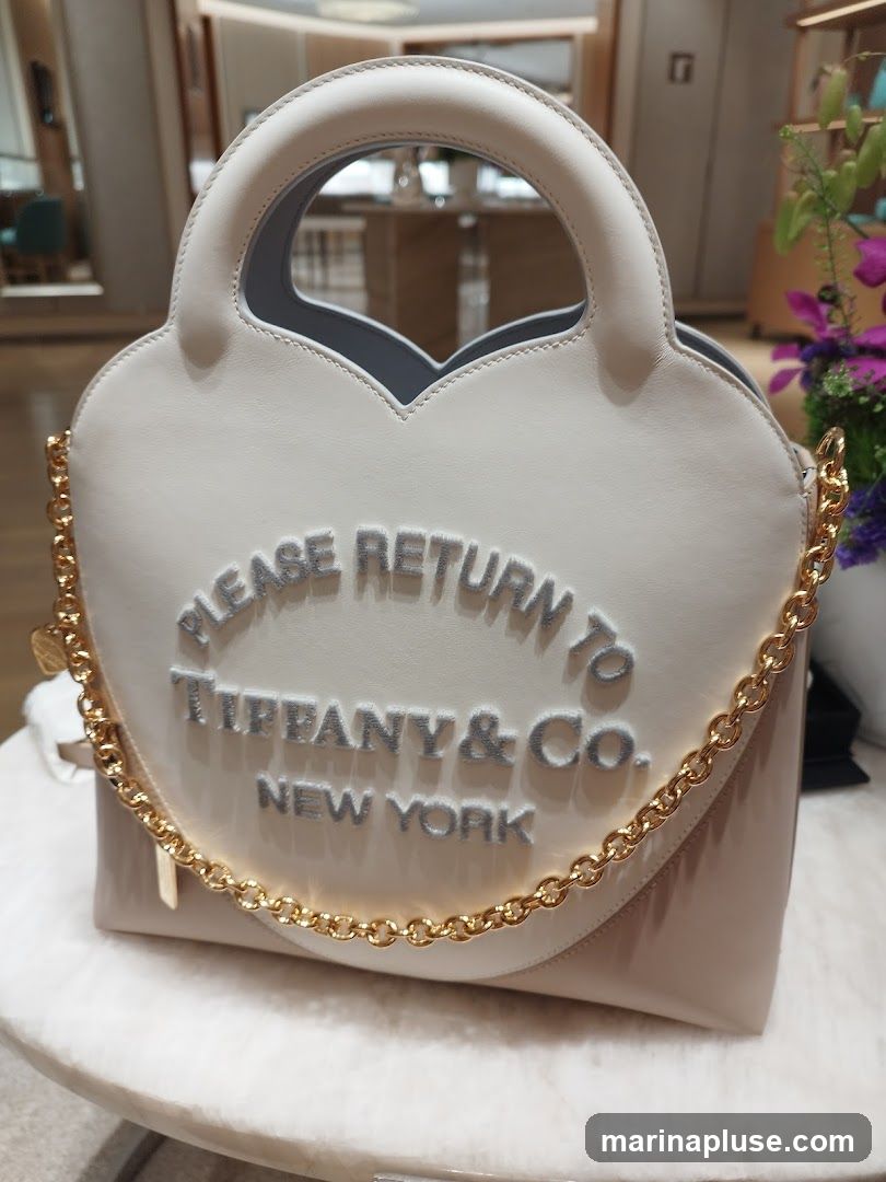 A light-colored, heart-shaped handbag with a gold chain strap is displayed on a marble surface. The bag features embossed text that reads "PLEASE RETURN TO TIFFANY & Co. NEW YORK" in a light blue color. The background appears to be an interior of a store with blurred displays and lighting.