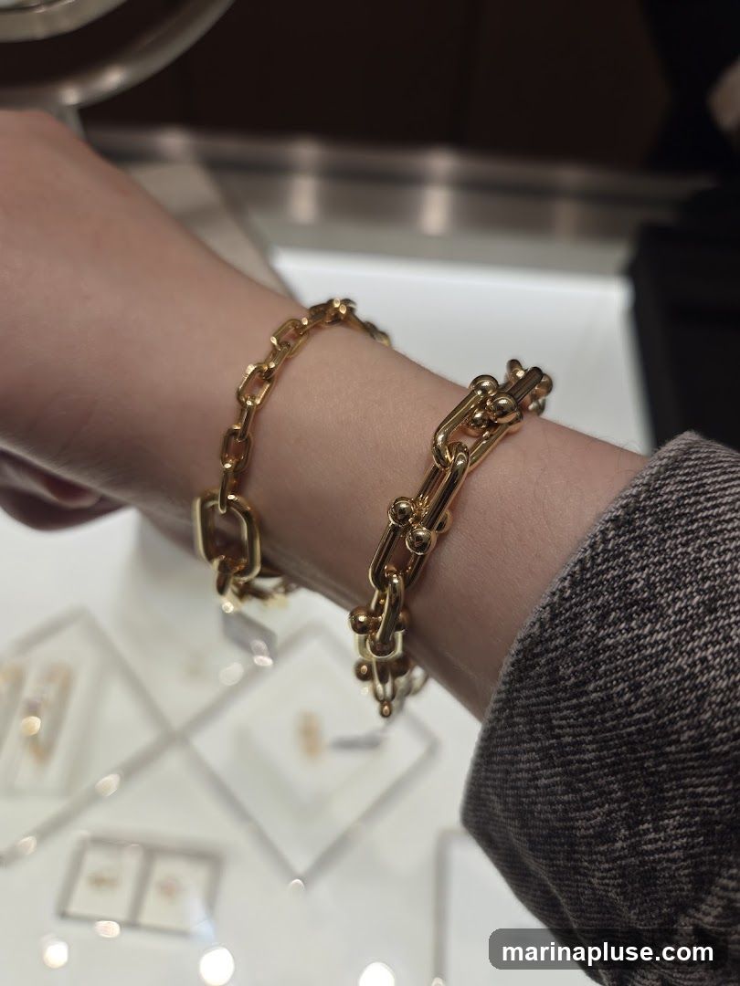 A close-up shot of a person's wrist wearing two gold chain bracelets. The bracelets are made of interlocking oval links and some links have small gold spheres attached. In the background, out of focus, are display cases with jewelry.