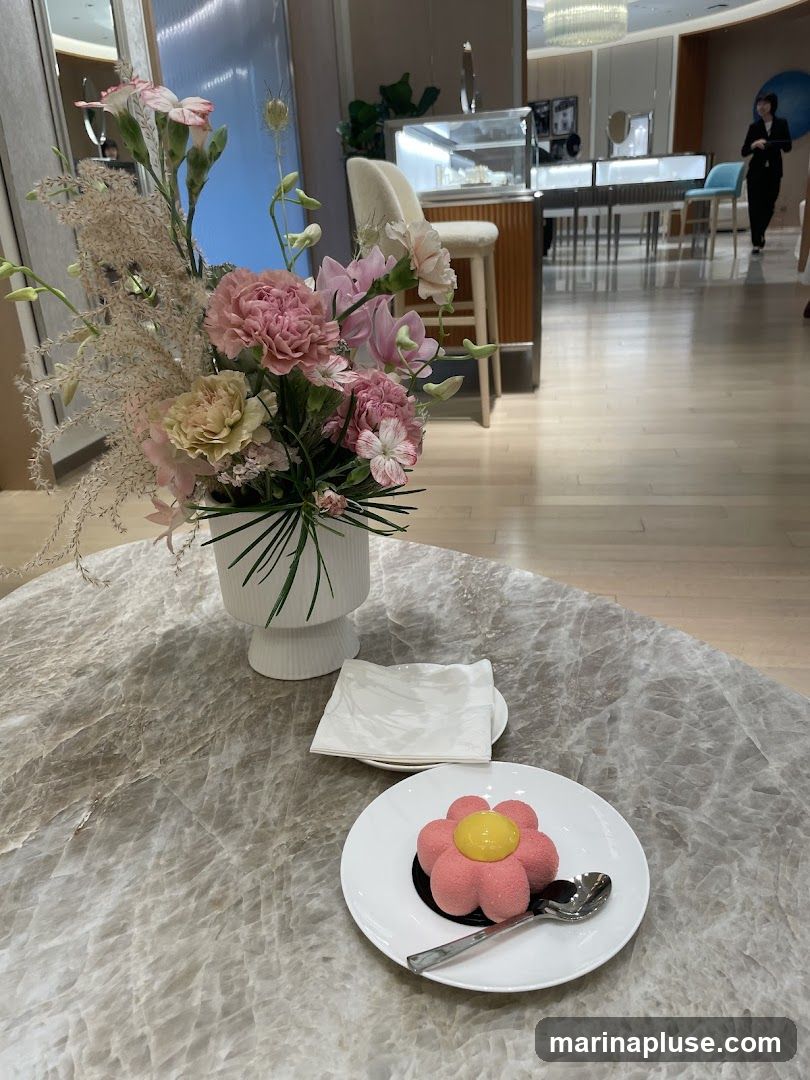 A pink flower-shaped dessert with a yellow center sits on a white plate with a spoon on a marble table. A vase of pink and white flowers is to the left. In the background, a store interior with display cases and seating is visible.