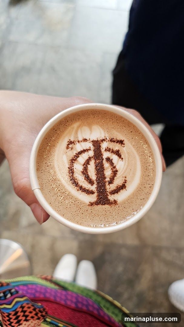 A hand holds a white paper cup filled with a latte. The latte art on top is a dark brown logo, possibly a stylized 'TC' or 'TP'. The background is blurred, showing a floor and the legs of a person wearing white shoes and a colorful patterned skirt.