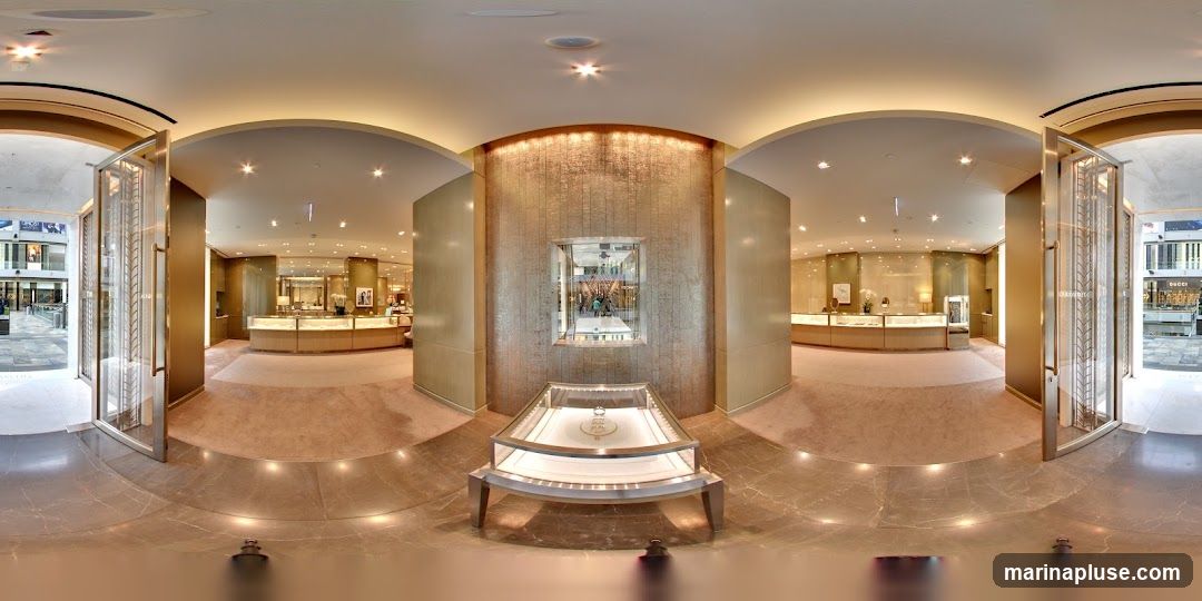 A panoramic view of the interior of a high-end jewelry store. The entrance features large glass doors with ornate metal detailing. Inside, the store is spacious and elegantly designed with soft lighting. Display cases with jewelry are arranged around the perimeter, and a central display case showcases a prominent item. The walls are adorned with textured panels and subtle lighting. Through the entrance, an outdoor shopping area is visible with other storefronts.