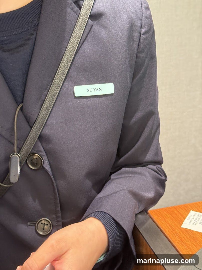 A close-up shot shows a person wearing a dark blue blazer. A light blue name tag with the name "SU YAN" printed in black is pinned to the lapel. The person is also wearing a dark sweater underneath the blazer, and a black strap from a bag is visible across their chest. Part of a wooden surface and a small white slip of paper are visible in the lower right corner.