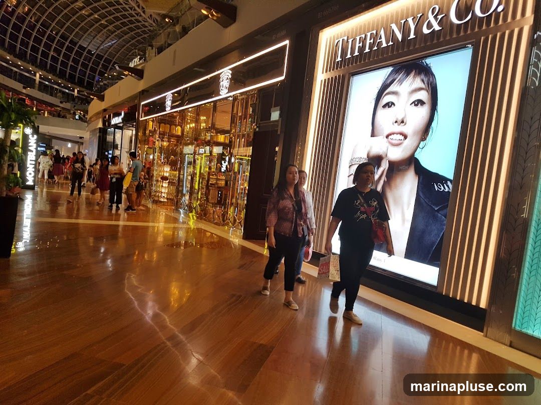 A wide shot of a polished wooden floor in a mall, reflecting the bright lights of the surrounding shops. On the right, a large advertisement for Tiffany & Co. features a close-up of a woman's face. To the left, a Montblanc store is visible, along with other shops and shoppers walking by. The ceiling features a modern, arched glass design.