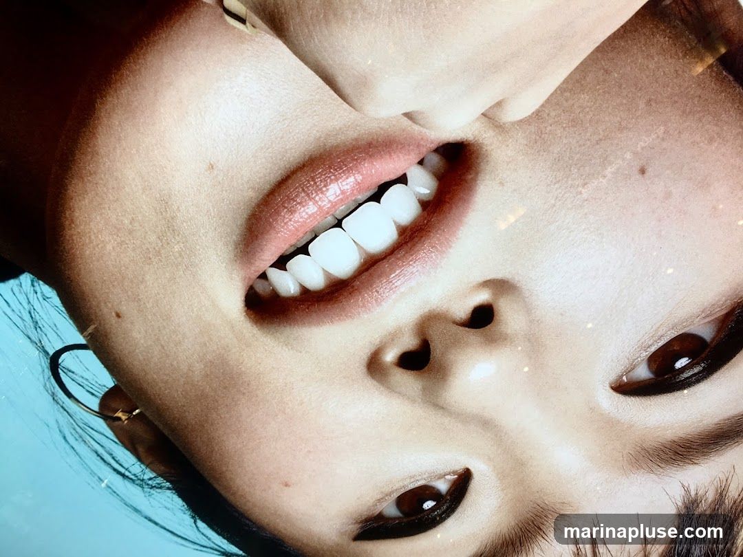 A close-up, upside-down view of a person's face, focusing on their mouth, teeth, and eyes. The person has full lips with a glossy pink lipstick and bright white teeth. Their eyes are dark and appear to have dramatic eyeliner. The background is a solid light blue.