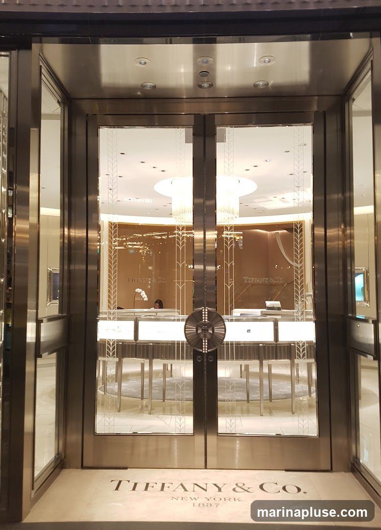 A view of the entrance to a Tiffany & Co. store. The entrance features double glass doors with decorative etching. The Tiffany & Co. logo and "NEW YORK 1837" are visible on the floor mat. Inside, display cases with jewelry are illuminated, and a chandelier hangs from the ceiling.
