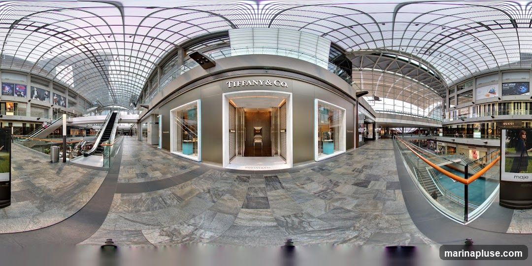A panoramic view of the interior of a modern shopping mall, with a prominent Tiffany & Co. store as the central focus. The store features a large, ornate entrance with double doors and illuminated display windows on either side showcasing jewelry. The Tiffany & Co. logo is visible above the entrance and on the floor mat. Other stores, escalators, and architectural elements of the mall are visible in the background.
