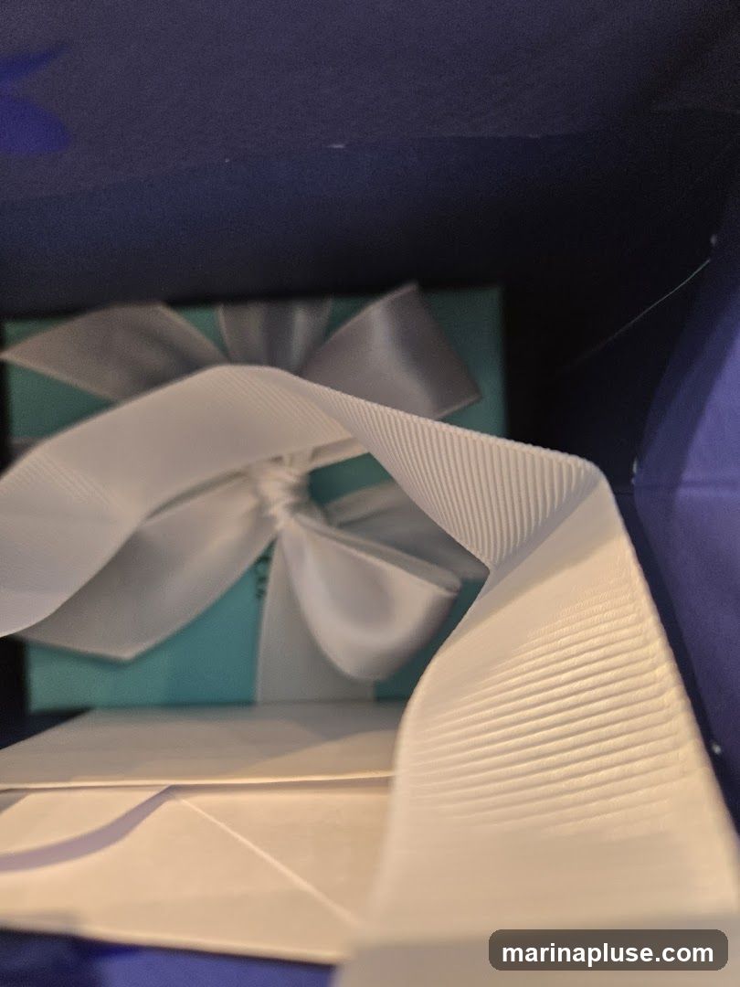 A close-up view inside a dark blue box, revealing a turquoise gift box tied with a white ribbon. The ribbon is prominently featured in the foreground, with its textured surface visible. A white envelope or paper is also visible beneath the gift box.