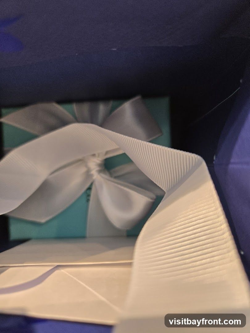 A close-up view inside a dark blue box, revealing a turquoise gift box tied with a white ribbon. The ribbon is prominently featured in the foreground, with its textured surface visible. A white envelope or paper is also visible beneath the gift box.