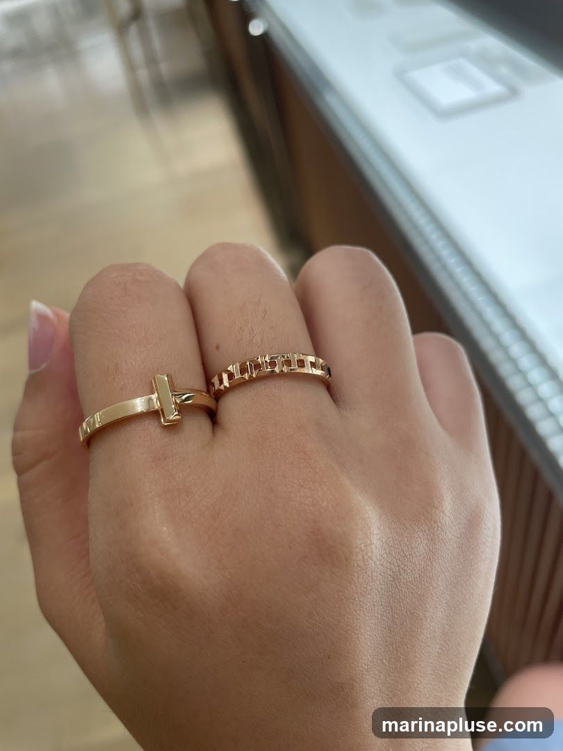 A close-up shot of a hand wearing two gold rings. One ring is a simple band with a T-shaped charm. The other ring is a band with a repeating geometric pattern. The background appears to be a display case in a jewelry store.
