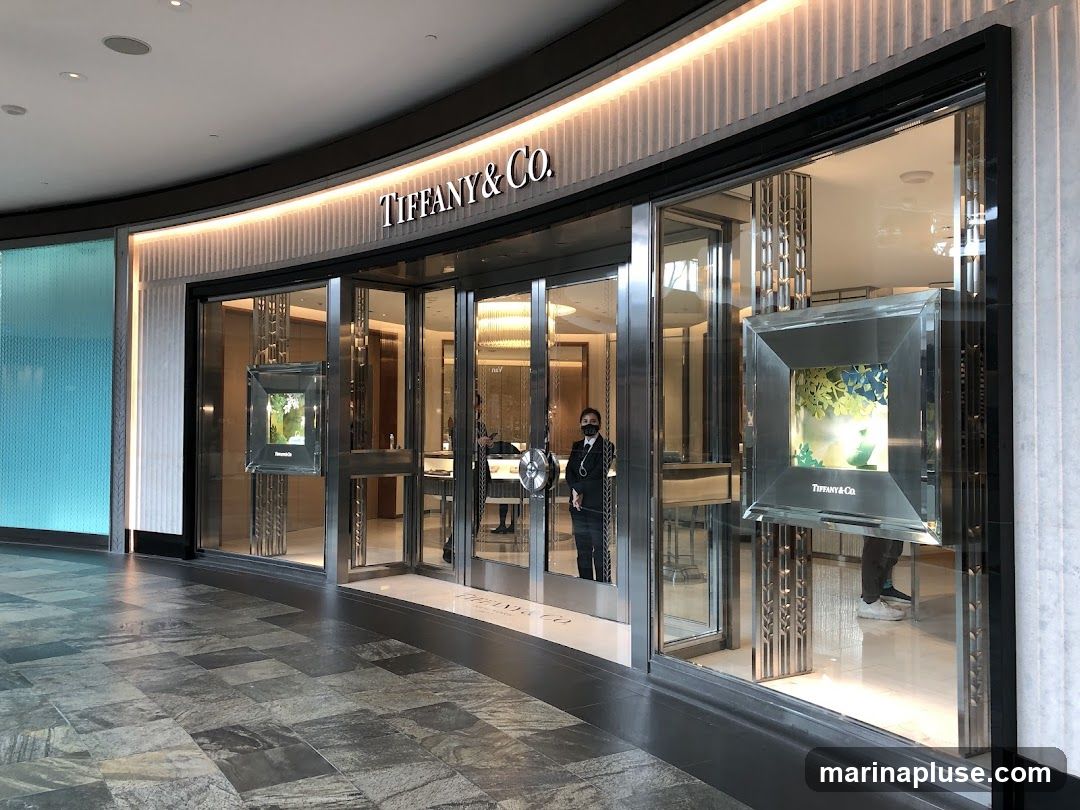 The exterior of a Tiffany & Co. store with large glass windows and doors. The store's name is prominently displayed in large letters above the entrance. Inside, displays of jewelry and a chandelier are visible. A staff member in a black suit and mask stands near the entrance.