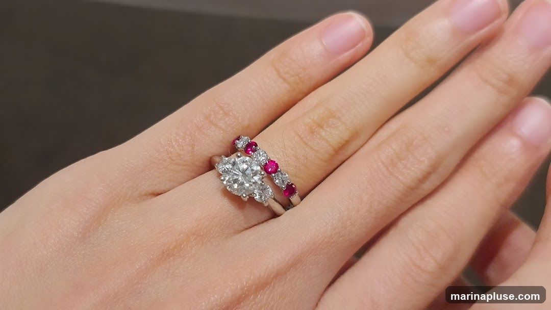 A close-up shot of a hand wearing two rings. The ring on the bottom features a large, round-cut diamond center stone, flanked by smaller diamonds. The ring on top is a band set with a row of alternating diamonds and round, red gemstones, likely rubies.