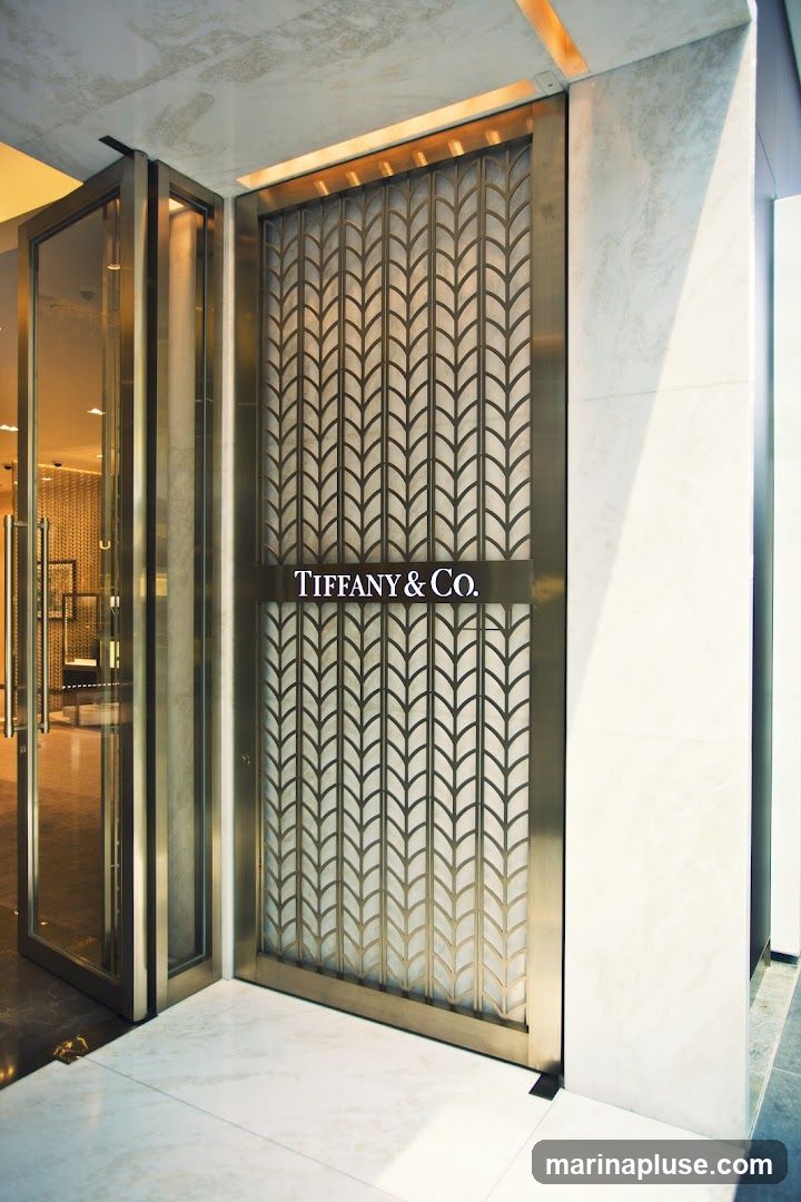 The entrance to a Tiffany & Co. store features a large, ornate metal gate with a repeating leaf pattern. A illuminated sign with the "TIFFANY & CO." logo is centered on the gate. To the left, a glass door and window reveal a glimpse of the store's interior, with displays and lighting visible. The floor is made of polished marble tiles.