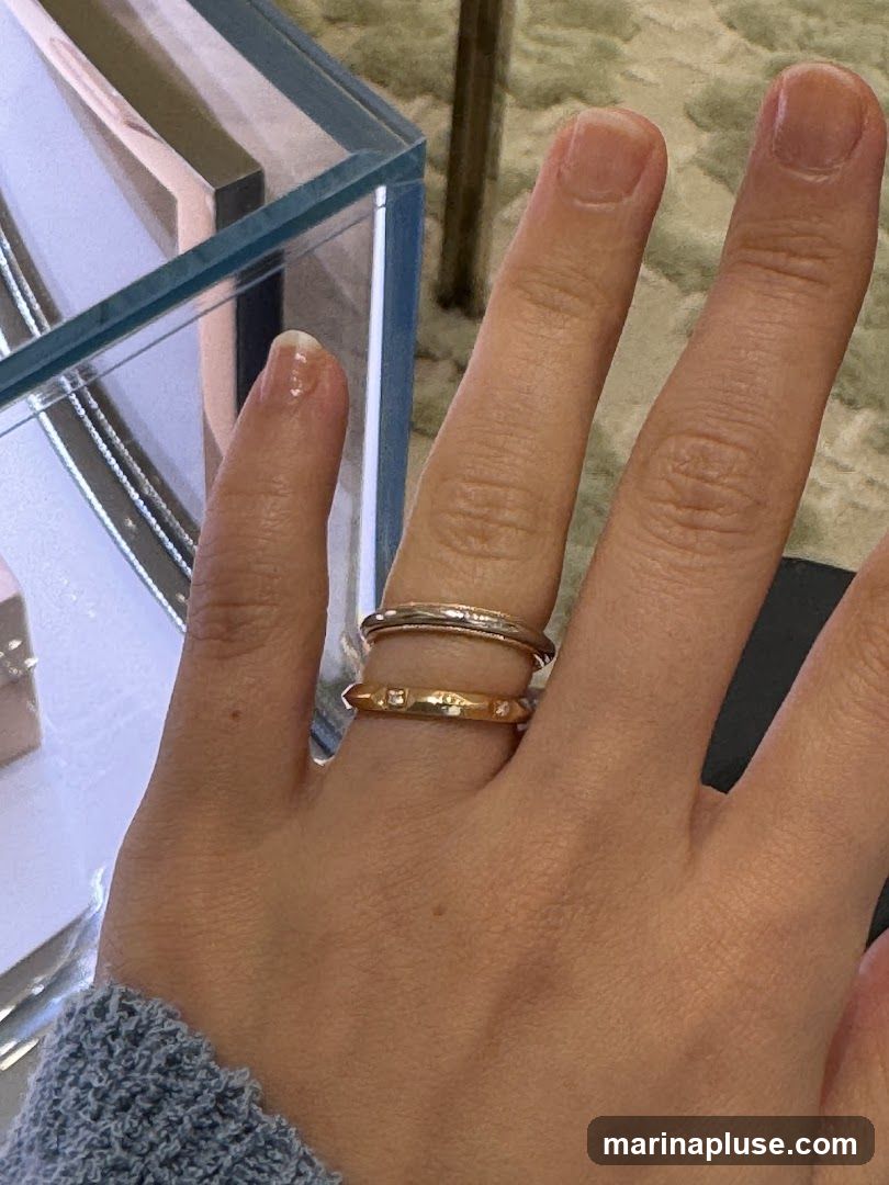 A close-up shot of a person's hand wearing two rings. The top ring is silver and thin, while the bottom ring is gold and features small, sparkling stones. The hand is resting on a textured surface, possibly a carpet or fabric. In the background, a glass display case is visible, suggesting the rings might be in a jewelry store.