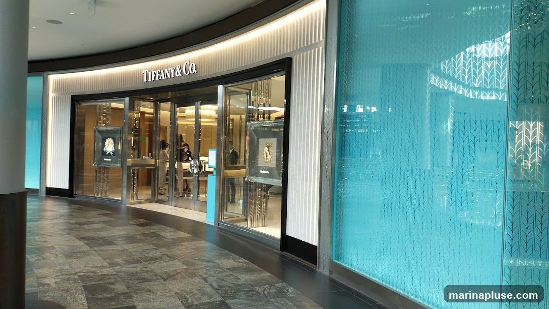 The exterior of a Tiffany & Co. store. The store has a large glass facade with the Tiffany & Co. logo visible above the entrance. Inside, displays of jewelry can be seen. The store is decorated with vertical ribbed panels and a large turquoise glass panel with a repeating leaf pattern.