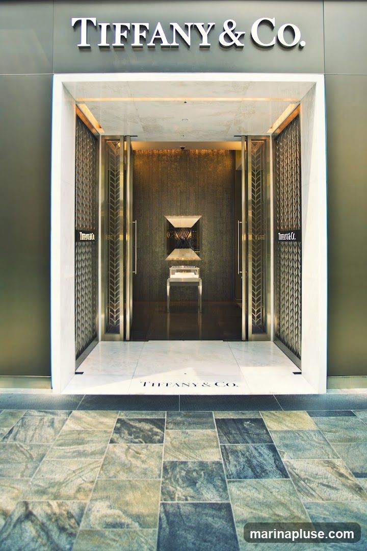 The exterior of a Tiffany & Co. store. The entrance features large glass doors with ornate metal detailing. Above the entrance, the Tiffany & Co. logo is prominently displayed in silver lettering. The floor inside the entrance is marble, with the Tiffany & Co. logo also visible on the floor. The exterior facade is a dark, muted color, contrasting with the bright interior visible through the doors.