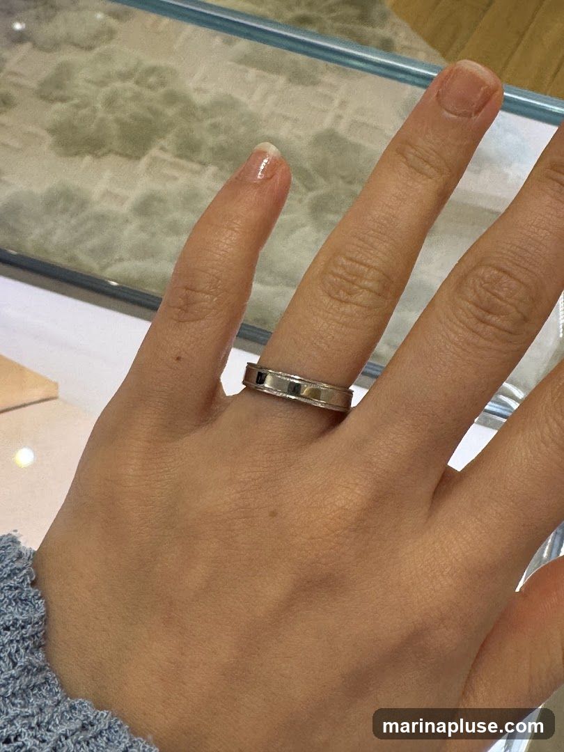 A close-up shot of a hand wearing a silver ring. The ring has a textured band with a polished center. The background appears to be a display case, possibly in a jewelry store, with a blurred, marbled pattern visible.