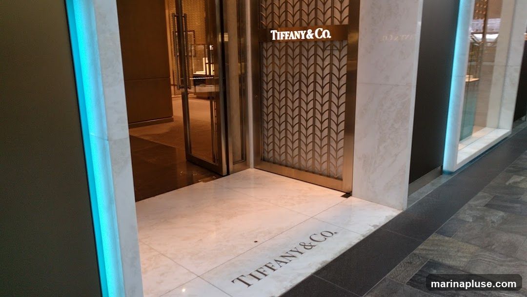 The entrance to a Tiffany & Co. store. The store's name is visible on a sign above the door and also on the floor in front of the entrance. The entrance features a decorative metal screen and marble accents.