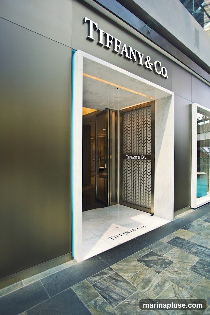 The exterior of a Tiffany & Co. store. The entrance is framed by white marble and features a decorative metal screen with the Tiffany & Co. logo. The store's name is also visible in large silver lettering above the entrance. The floor inside the entrance and leading up to it is tiled with marble, with the Tiffany & Co. logo subtly incorporated into the design on the floor. The building facade is dark gray, with a thin turquoise accent line.
