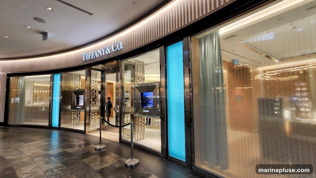 The exterior of a Tiffany & Co. jewelry store. The store has a curved facade with large glass windows and doors. The Tiffany & Co. logo is prominently displayed above the entrance. Inside, displays of jewelry and lighting fixtures are visible. The store is decorated in a luxurious style with metallic accents and soft lighting.