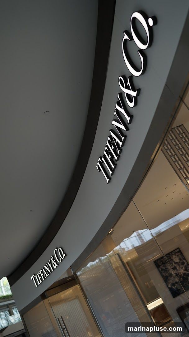 A close-up, angled view of the exterior signage and entrance of a Tiffany & Co. store. The iconic Tiffany & Co. logo is prominently displayed in illuminated white lettering on a dark gray background above a glass storefront. The reflection on the glass shows interior lighting and decor.