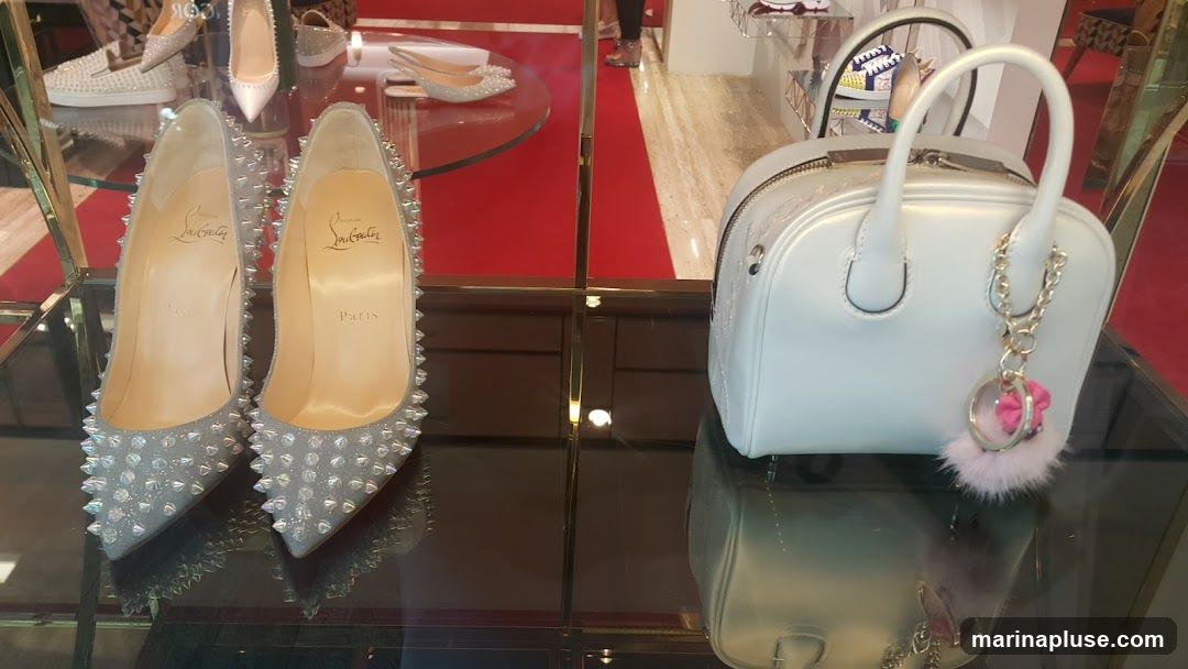 A display of luxury footwear and a handbag. In the foreground, two silver, spike-embellished high-heeled shoes with the Christian Louboutin logo and "Paris" visible on the insoles are showcased. To the right, a white, structured handbag with a gold chain and a fluffy pink pom-pom charm is displayed. In the background, other shoes and accessories are visible on shelves, with a red carpeted floor adding to the opulent presentation.