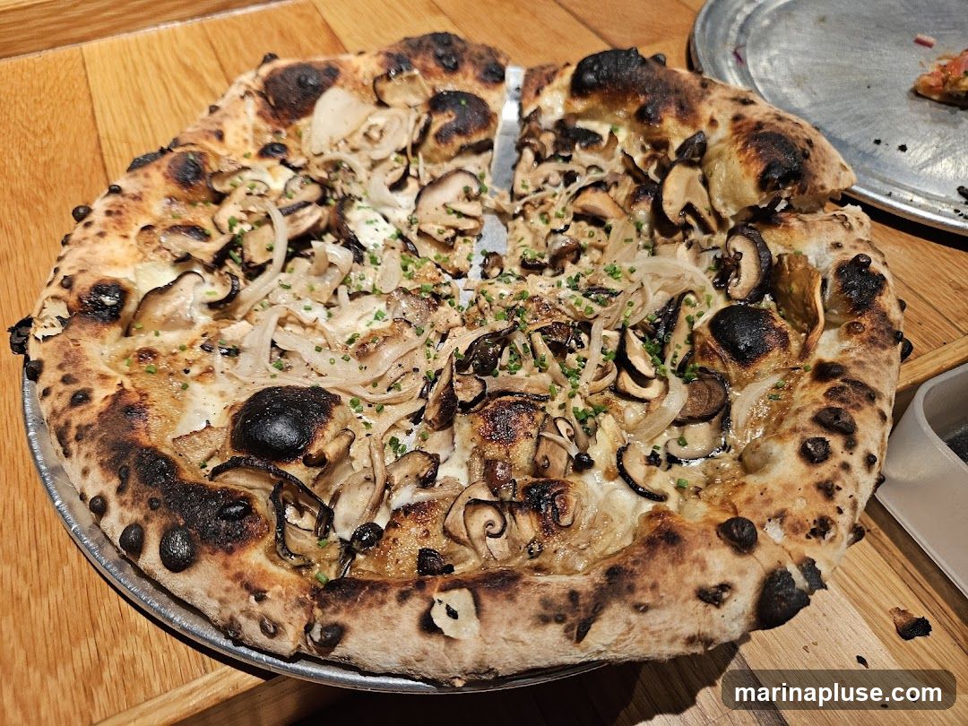 A close-up shot of a whole mushroom pizza with a charred crust, topped with various mushrooms, onions, and chives. The pizza is served on a metal pan on a wooden table. A slice of pizza is visible in the background on another pan.