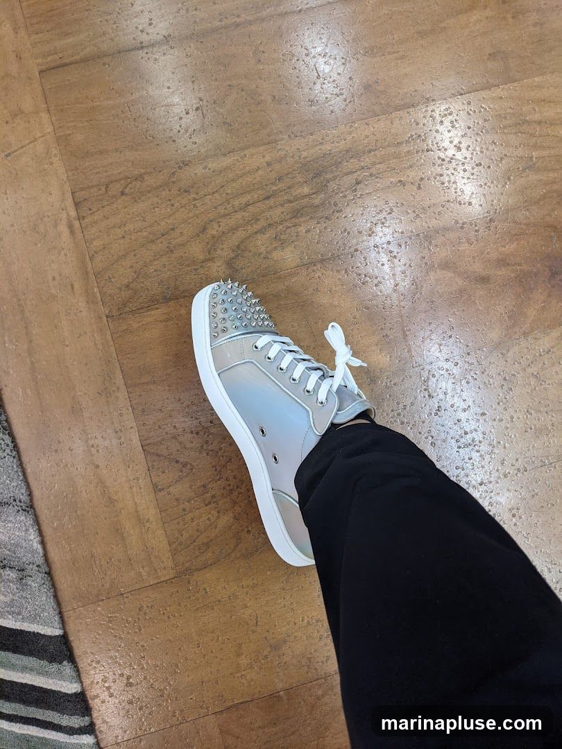 A person is wearing a pair of silver, high-top sneakers adorned with silver spikes on the toe box. The sneakers have white laces and white soles. The person is also wearing black pants. The shoes are on a wooden floor.