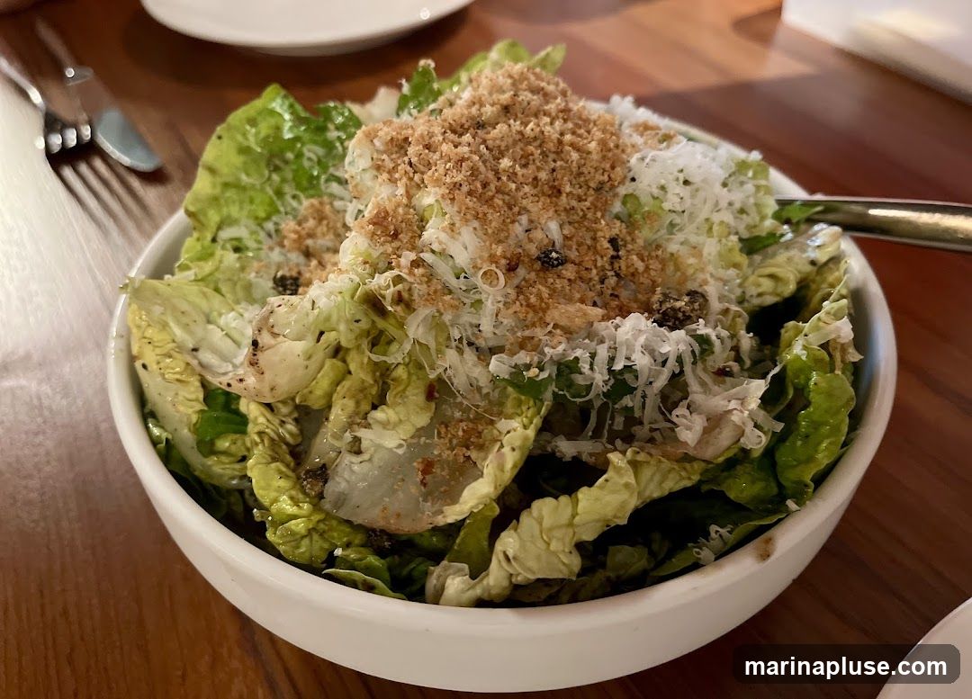 A close-up shot of a Caesar salad in a white bowl. The salad is made with romaine lettuce, topped with shredded parmesan cheese, croutons, and a dressing. A fork and knife are visible on the left side of the image.