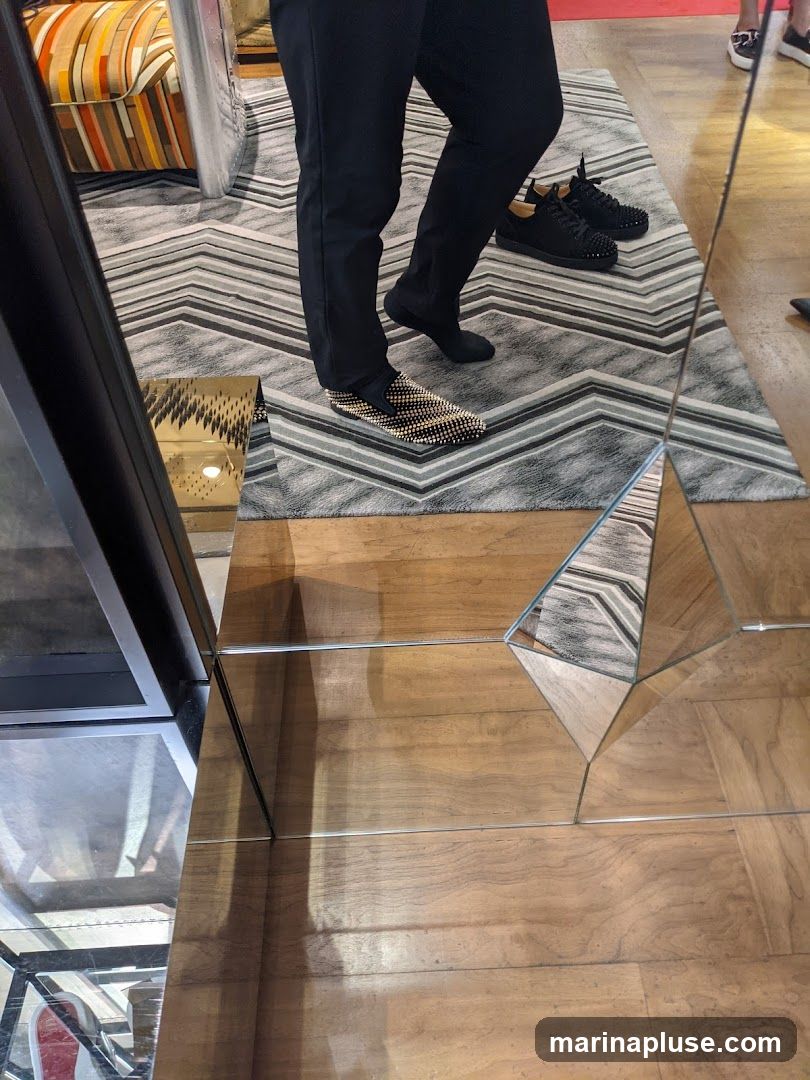 A person is standing on a rug in front of a mirror, trying on a pair of gold and black studded loafers. Another pair of black studded sneakers is visible on the rug behind them. The floor is made of wood, and there are mirrored geometric shapes on the floor. A colorful striped armchair is reflected in the mirror.