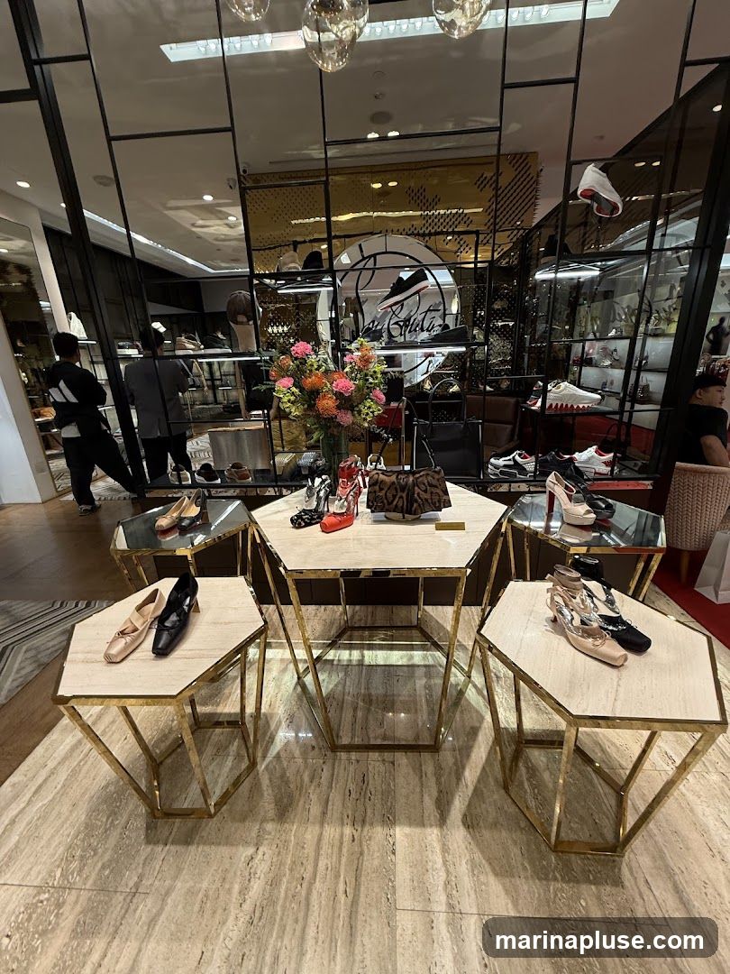 A well-lit interior of a high-end shoe store. In the foreground, hexagonal marble-topped tables with gold geometric bases display various women's shoes and handbags. A central table features red and black high heels and a leopard-print clutch. Other tables showcase nude ballet flats, black pumps, and embellished heels. In the background, shelves are stocked with more footwear, and a circular logo with a shoe silhouette is visible on the wall. Two individuals are browsing in the background.