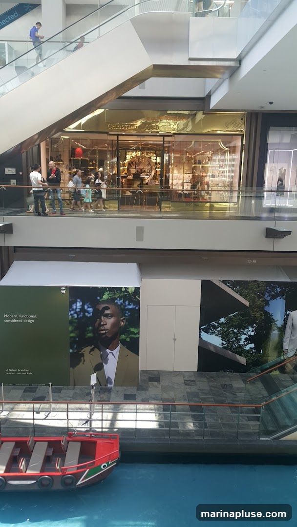 An interior view of a multi-level shopping mall. On the upper level, a storefront with a gold facade and the name "CHRISTIAN LOUBOUTIN" is visible. People are seen walking past the store. Below this, large advertisements are displayed, one featuring a portrait of a man with the text "Modern, functional, considered design" and "A fashion brand for women, men and kids." Another advertisement shows a man in a suit. In the foreground, a red boat with seating is on a blue water channel. Escalators and railings are also visible throughout the space.