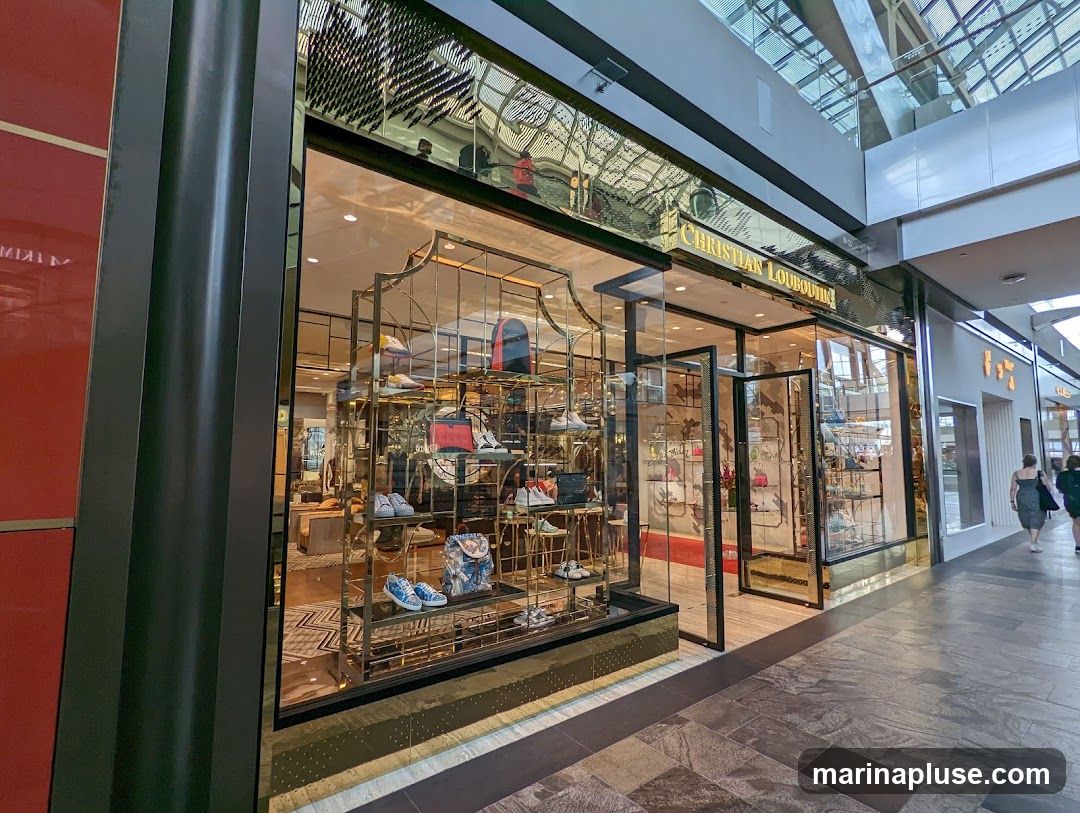 The exterior of a Christian Louboutin store, featuring a large glass window display showcasing various shoes and handbags on gold shelves. The store's name is prominently displayed in gold lettering above the entrance. The entrance has glass doors and a gold frame. The store is located within a modern shopping mall with a glass ceiling.