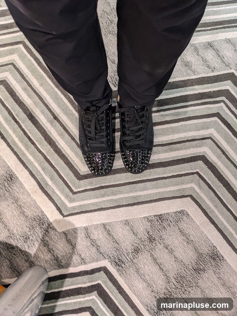 A pair of black lace-up sneakers with silver studs on the toe box, worn with black pants, resting on a chevron-patterned rug.
