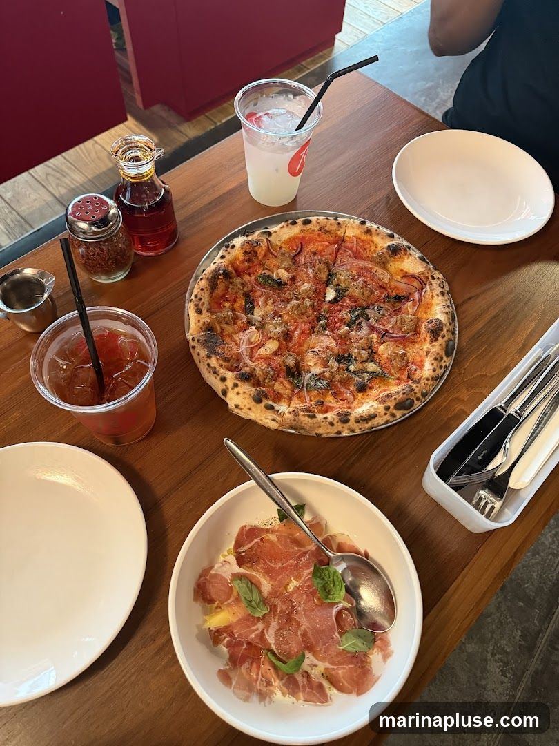 A table setting with a pizza, a bowl of prosciutto and cheese, two drinks, and condiments. The pizza is topped with sausage, onions, and herbs. The appetizer appears to be thinly sliced cured ham over a creamy base, garnished with basil. One drink is a clear beverage with ice and a straw, served in a plastic cup with a red logo. The other is a darker iced tea. A shaker of red pepper flakes and a small bottle of oil are also on the table, along with cutlery.