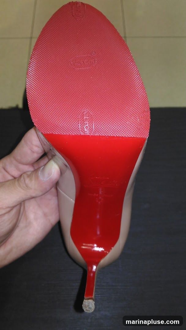 A close-up view of the sole of a high-heeled shoe, featuring the iconic red Christian Louboutin sole. The sole is textured and has several embossed logos, including "Vibram". The heel is slender and also red. A hand is holding the shoe from the side.