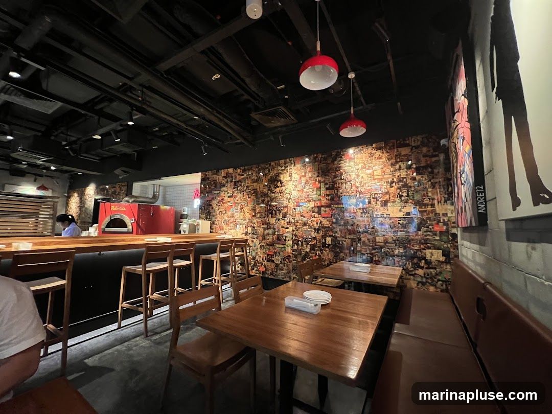 The interior of a restaurant features a long wooden bar with stools, a red pizza oven, and a wall covered in a collage of vintage images. Tables and booth seating are visible, along with modern pendant lights and exposed ceiling infrastructure. A person is working behind the bar.