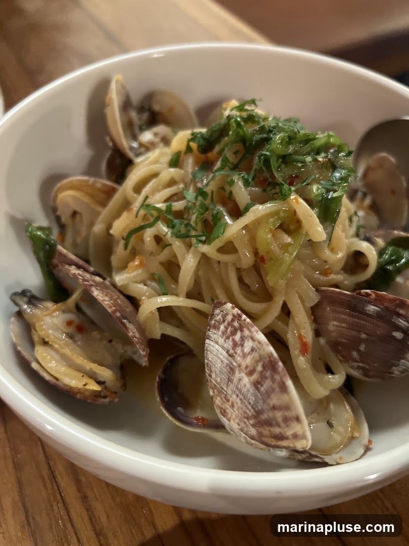 A close-up shot of a white bowl filled with linguine pasta and clams. The pasta is tossed in a light sauce and garnished with chopped green herbs. Several clams are visible, some open and some closed, nestled among the pasta. The dish is presented on a wooden surface.