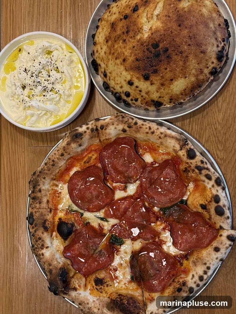 A close-up overhead shot of a pepperoni pizza, a round of bread, and a bowl of dip. The pizza has a charred crust and is topped with melted cheese, tomato sauce, and slices of pepperoni. The bread is also well-baked with a dark, blistered crust. The dip appears to be a creamy white substance, possibly ricotta or burrata, drizzled with olive oil and sprinkled with black pepper.