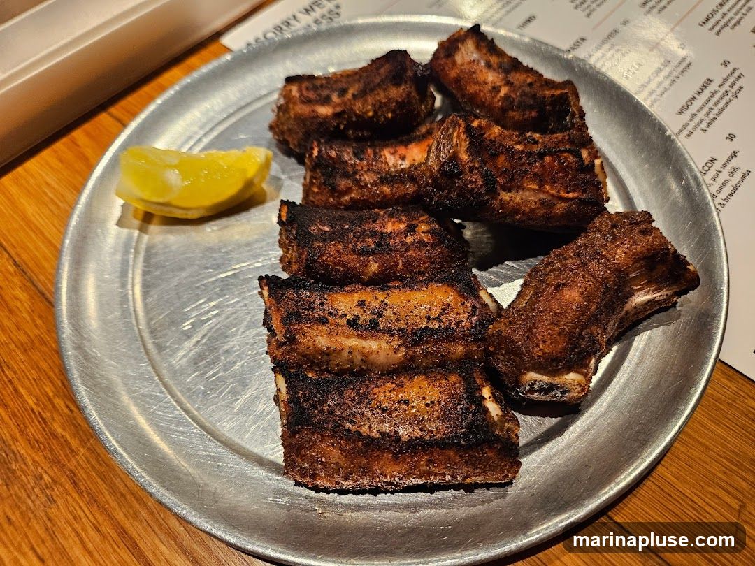 A metal plate holds several pieces of seasoned and grilled ribs, with a lemon wedge on the side. A menu is partially visible in the background.